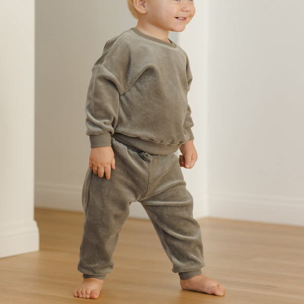 A young child stands barefoot on a wooden floor, smiling and cozy in the Velour Relaxed Sweatpant set with matching long-sleeve sweatshirt.