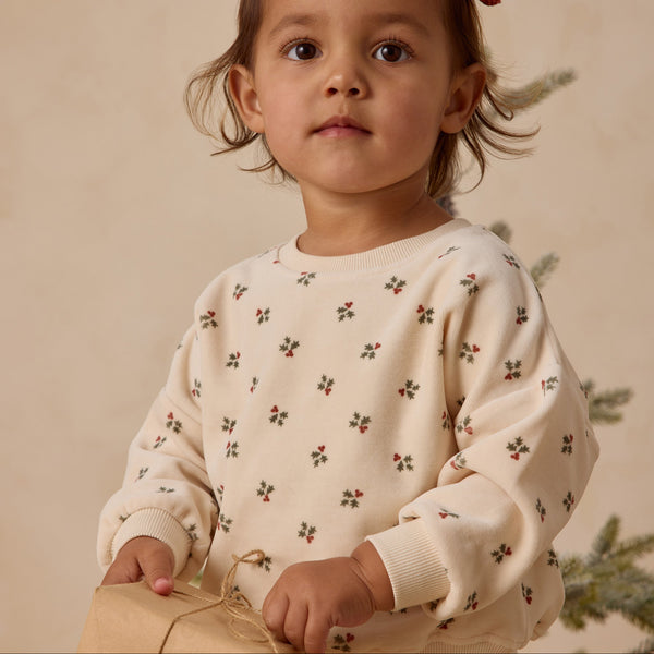 A young child wears the Velour Relaxed Sweatshirt (2 styles), featuring small holly berry prints, and stands indoors holding a wrapped gift. A blurred evergreen branch appears in the background.