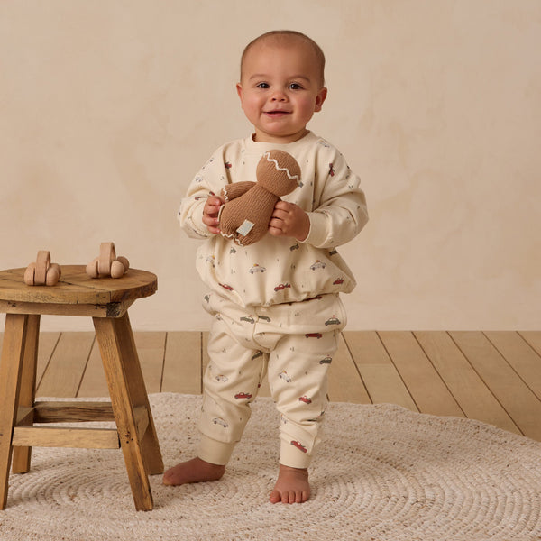 A smiling baby in a Relaxed Fleece Sweatshirt stands on a textured rug holding a brown stuffed toy, with a wooden stool beside them displaying two wooden toy cars—all set on a wooden floor.