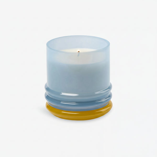 The 7oz Stacked Glass Candle, made with white soy wax, glows in a translucent, multi-colored glass holder with a rounded base featuring yellow and blue accents, set against a plain white background.