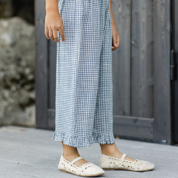 A child in Wide Leg Ruffle Pant and cream crochet shoes stands on a gray wood floor by a dark wood door.