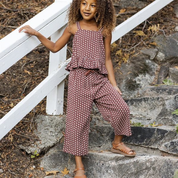 A young girl with curly hair stands on outdoor stone steps, smiling and holding a white railing while wearing the Kayli Set - Chocolate Polka Dot and brown sandals.
