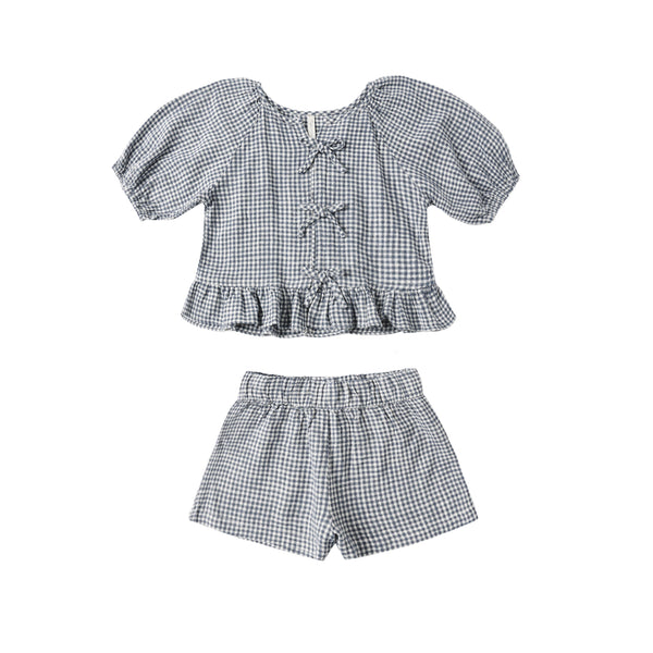 The Juniper Set is a children’s outfit with a blue and white marine gingham short-sleeve top featuring ruffled hem and bow details, paired with matching elastic-waist linen shorts, shown on a white background.