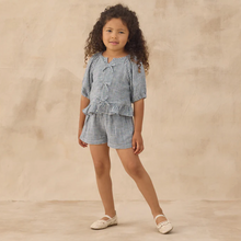Load image into Gallery viewer, A young child with long curly hair stands against a beige textured backdrop, wearing the Juniper Set in marine gingham with ruffle detail and matching linen shorts, paired with cream shoes.