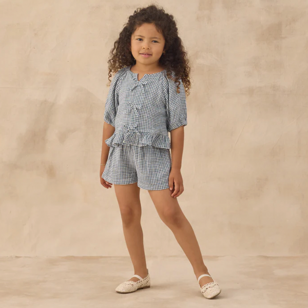 A young child with long curly hair stands against a beige textured backdrop, wearing the Juniper Set in marine gingham with ruffle detail and matching linen shorts, paired with cream shoes.
