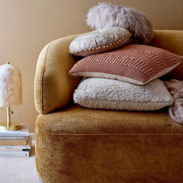 A mustard-yellow velvet sofa features a 20" Cream Boucle Pillow and three textured throws stacked on one end, beside a small marble table lamp and books against a beige wall.