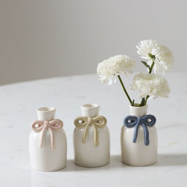 The Budvase w/ Bow set features three small white ceramic vases with pastel pink, yellow, and blue bows—perfect for spring event decor. The vase with the blue bow displays white flowers while the others sit empty on a marble surface.