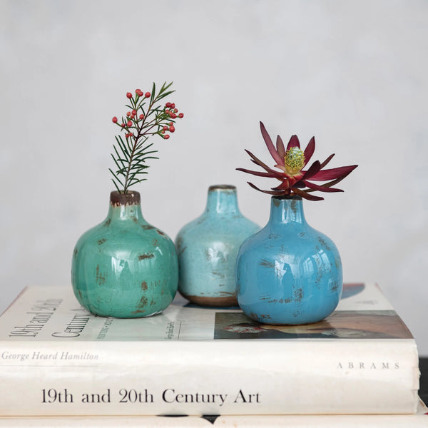 Three Terracotta Mini Vases in turquoise and blue, each with a single flower, are displayed on two stacked art books set against a light, neutral background for a vintage-inspired look.