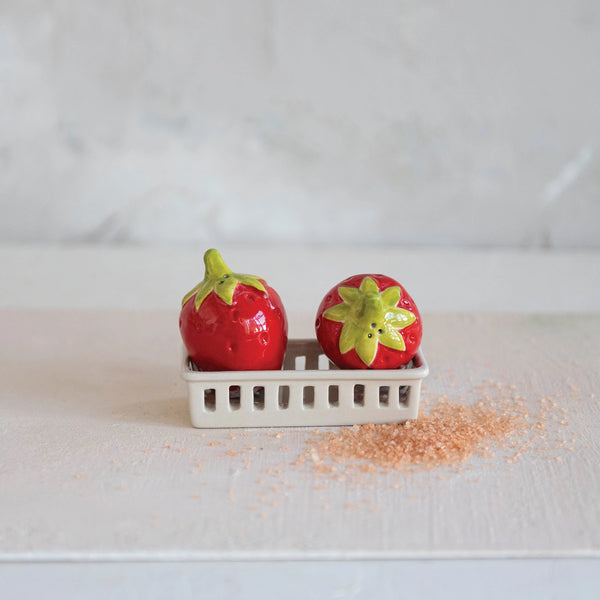 The Strawberry S+P Shakers, shaped like ceramic strawberries, rest in a small berry basket holder on a light surface with pink granules scattered nearby and a plain, light background.