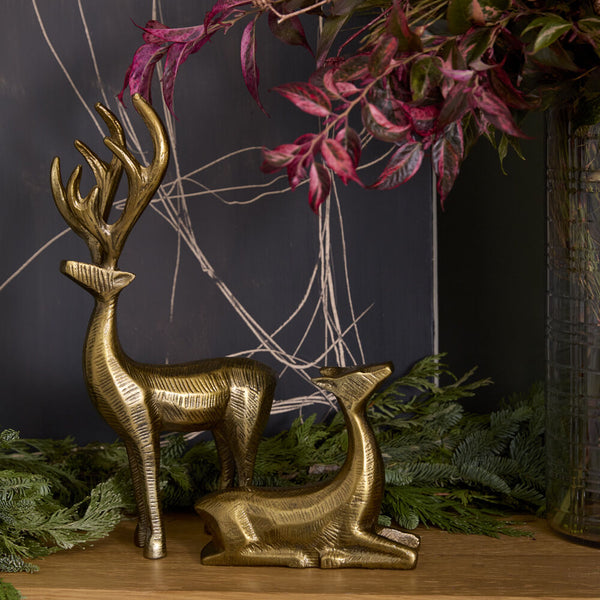 The Golden Prancer Reindeer Figurines, one standing and one lying down, are displayed among green foliage and pinkish leaves on a surface, with a dark background featuring subtle linear light patterns—ideal for festive décor.