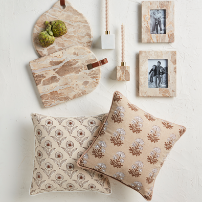 A flat lay featuring Mudpie Block Print Flower Knot Pillows in two styles, two marble cutting boards with artichokes, two marble photo frames with black-and-white photos, and marble-handled utensils on a white textured surface.