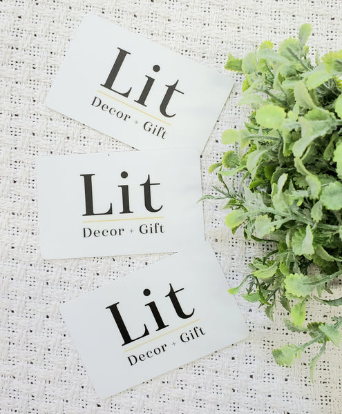 Three Gift Cards from Lit Decor and Gift are displayed on a textured white surface beside a small green plant—an ideal choice for gifting and redeeming unique decor treasures.