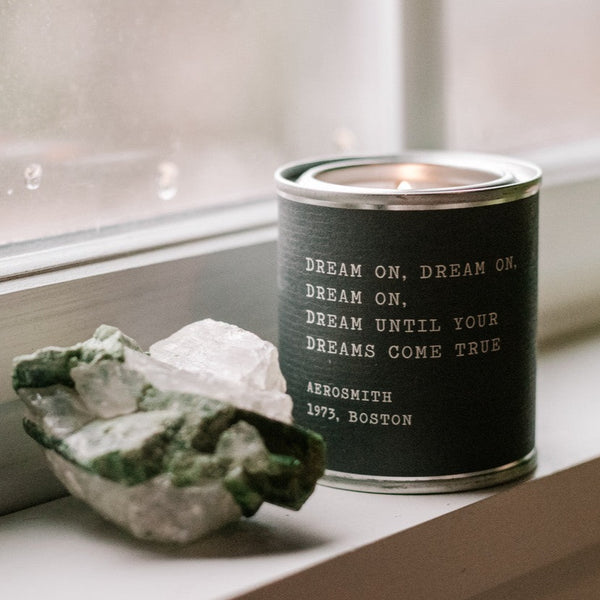 A Legends Candles - Pick From 20 Quotes candle by Lit Decor and Gift, made with soy wax and vegan-friendly, sits lit on a windowsill beside two unpolished crystals—one green, one clear.